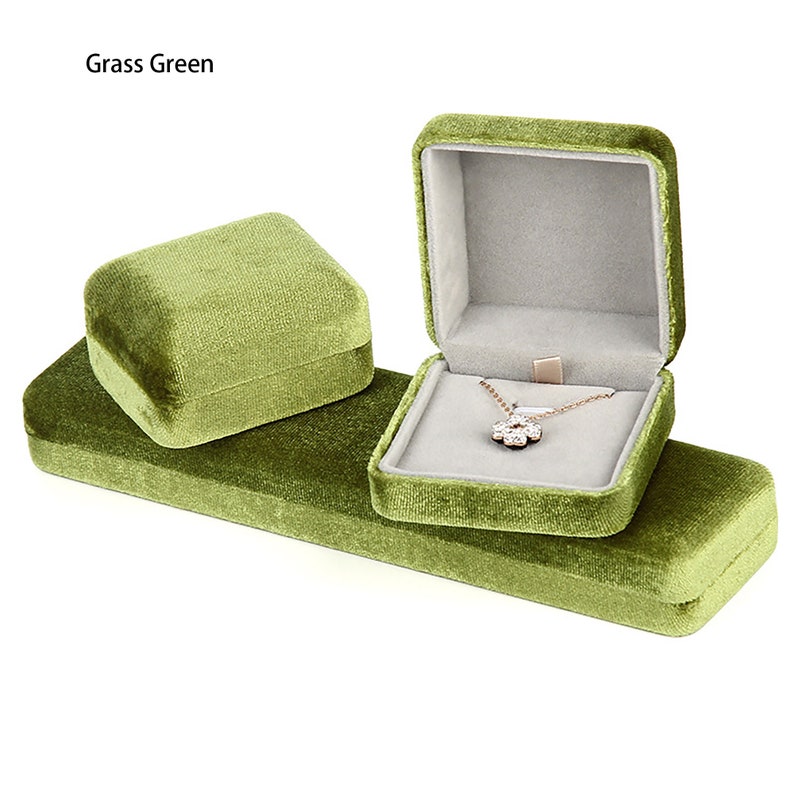 May include: A set of three green velvet jewelry boxes. The largest box is rectangular and has a smaller square box inside. The smallest box is square and is sitting on top of the larger box. The square box has a necklace with a flower pendant inside.