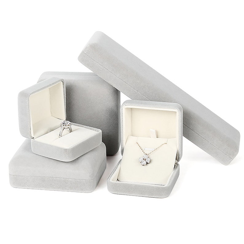May include: Set of four gray velvet jewelry boxes. Two boxes are square and have a white interior. One box is rectangular and has a white interior. One box is square and has a gray interior. The boxes are empty except for a ring in one box and a necklace in another box.