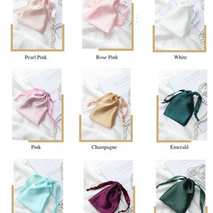 May include: Assortment of ten satin drawstring pouches in various colors, including pearl pink, rose pink, white, pink, champagne, emerald, fresh blue, purple, and deep green. The pouches are arranged in three rows of three, with each pouch sitting on a white surface with pearl beads scattered around it.