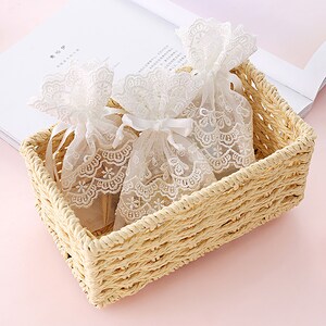 May include: Two white lace drawstring bags with white ribbon ties are in a woven wicker basket. The basket is light brown and has a rectangular shape.