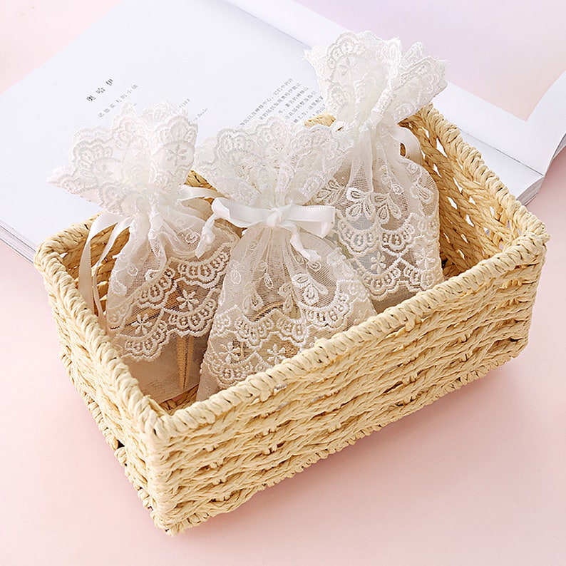 May include: Two white lace drawstring bags with white ribbon ties are in a woven wicker basket. The basket is light brown and has a rectangular shape.