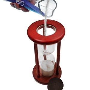 May include: A red wooden hourglass with a glass bulb filled with white sand. The sand is being poured from a clear glass bottle with blue sand.