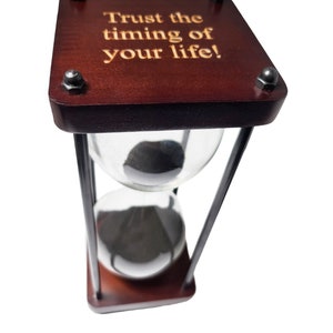 May include: A wooden hourglass with a black metal frame and a glass bulb. The top of the hourglass has a wooden plaque with the inscription "Trust the timing of your life!"