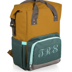 May include: A mustard and teal backpack with a grey handle and straps. The front pocket features the embroidered initials "TRS" in a script font. The backpack has a roll-top closure with a black buckle.