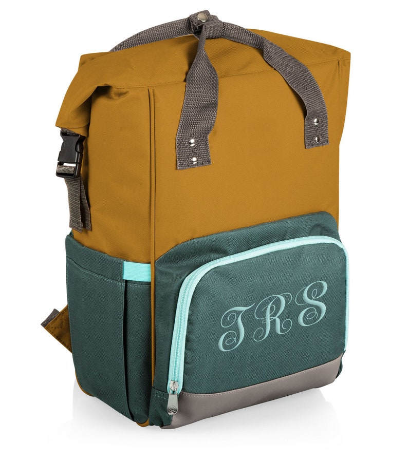 May include: A mustard and teal backpack with a grey handle and straps. The front pocket features the embroidered initials "TRS" in a script font. The backpack has a roll-top closure with a black buckle.