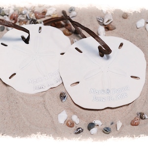 May include: Two white sand dollar ornaments with brown ribbon ties. The sand dollars are engraved with the text "Mark & Jenny June 10, 2016".