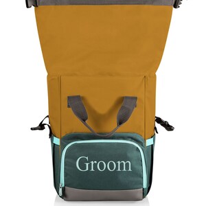 May include: A mustard yellow and teal backpack cooler with the word "Groom" embroidered on the front pocket. The cooler has a grey handle and trim, and a teal zip. It is designed to keep items cold.