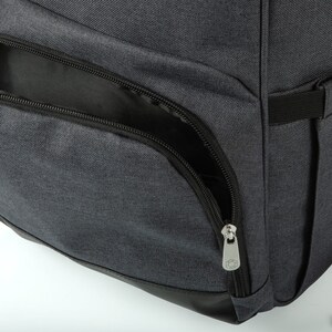 May include: Close-up of a gray backpack with a black zippered pocket and a black zipper pull. The zipper pull has a small hexagonal logo.