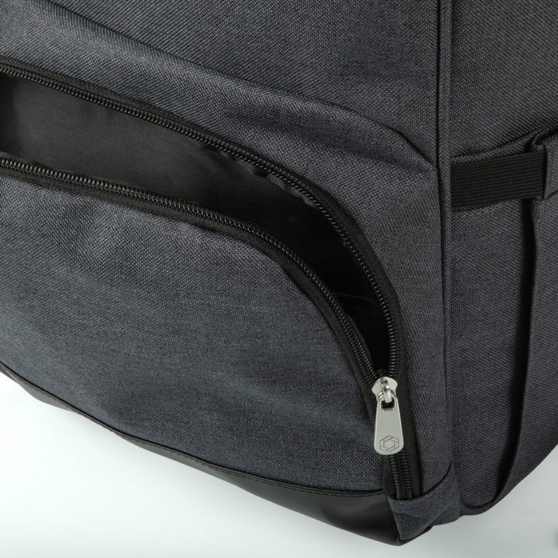 May include: Close-up of a gray backpack with a black zippered pocket and a black zipper pull. The zipper pull has a small hexagonal logo.