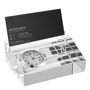 May include: A clear crystal desk organizer with a small clock and a business card holder. The clock has a white face with black Roman numeral hour markers and black hands. The business card holder is made of clear crystal and has a slot for holding business cards.