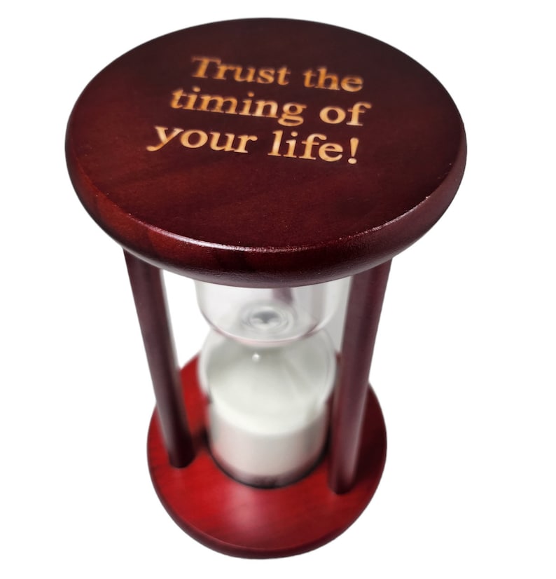 K&ouml;nnte beinhalten: Eine Sanduhr aus Holz mit roter Oberfl&auml;che und der Aufschrift "Trust the timing of your life!" auf der Oberseite.