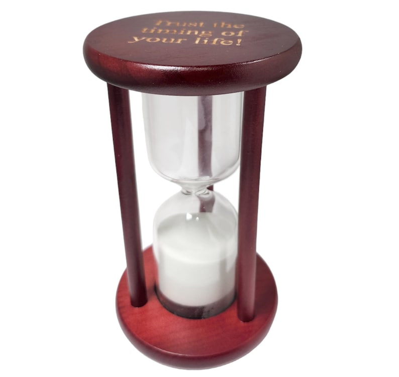K&ouml;nnte beinhalten: Eine Sanduhr aus Holz mit roter Oberfl&auml;che. Die Oberseite der Sanduhr tr&auml;gt die Inschrift "Trust the timing of your life!"