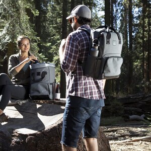 May include: Two people are in a forest setting. One person is sitting on a tree stump and holding a gray and black insulated cooler bag. The other person is standing and wearing a gray and black backpack cooler bag with a black water bottle in the side pocket.