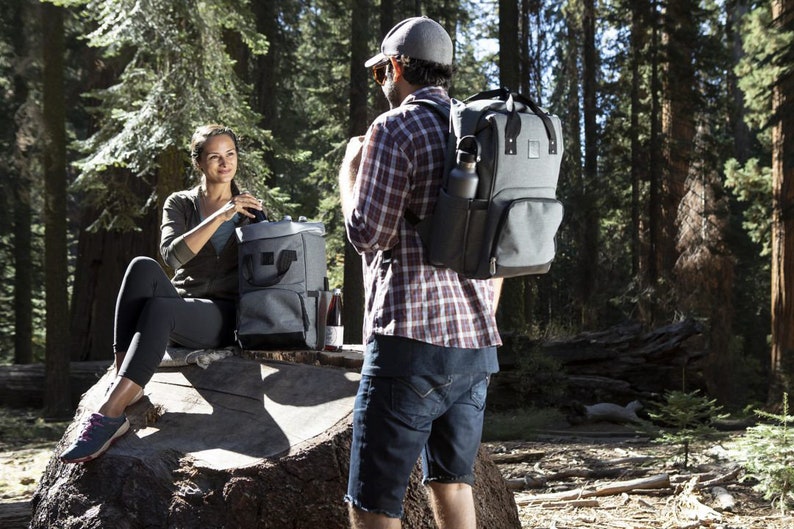 May include: Two people are in a forest setting. One person is sitting on a tree stump and holding a gray and black insulated cooler bag. The other person is standing and wearing a gray and black backpack cooler bag with a black water bottle in the side pocket.