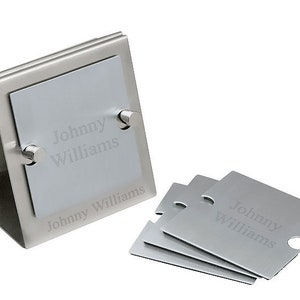 May include: A set of silver-colored metal coasters and a holder. The coasters and holder have the name "Johnny Williams" engraved on them. The holder is a triangular shape with a slot to hold the coasters. The coasters have a small cutout on one side.