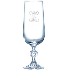 Op de afbeelding: Een heldere glazen champagnefluit met een decoratief ontwerp van kleine, heldere strasssteentjes in de vorm van een gestileerde werveling. De fluit heeft een ronde, gefacetteerde voet.