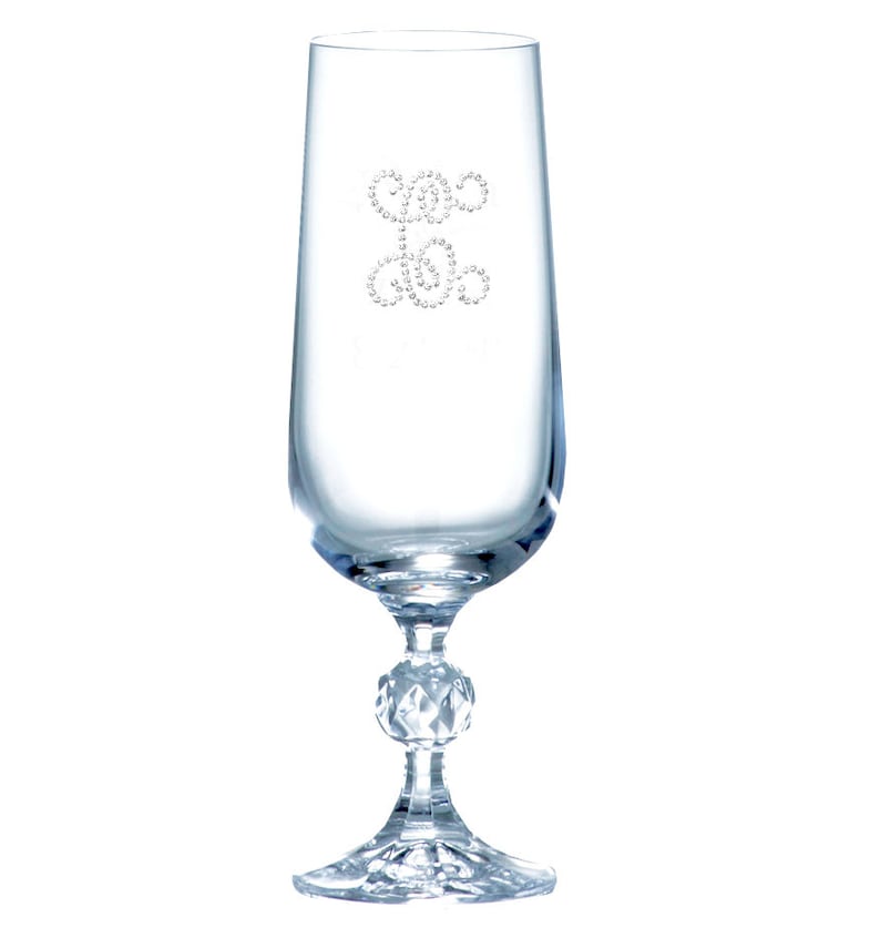 Op de afbeelding: Een heldere glazen champagnefluit met een decoratief ontwerp van kleine, heldere strasssteentjes in de vorm van een gestileerde werveling. De fluit heeft een ronde, gefacetteerde voet.