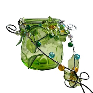 Pu&ograve; includere: Un barattolo di vetro verde con coperchio, adornato con un tralcio decorativo in filo metallico con foglie verdi, fiori trasparenti e perline colorate. Il barattolo ha una superficie strutturata ed &egrave; progettato per scopi decorativi.