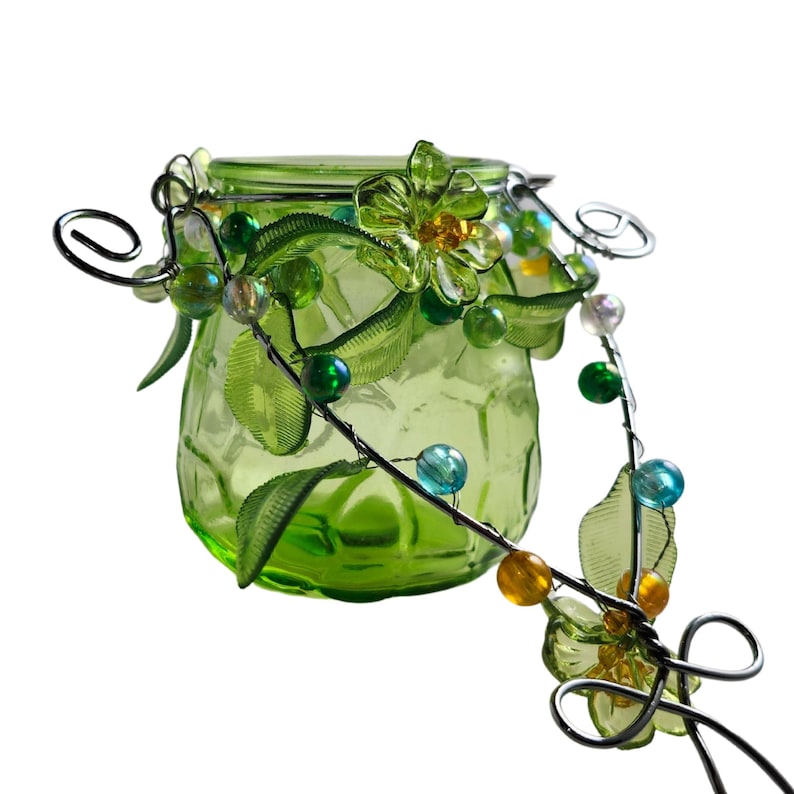 Pu&ograve; includere: Un barattolo di vetro verde con coperchio, adornato con un tralcio decorativo in filo metallico con foglie verdi, fiori trasparenti e perline colorate. Il barattolo ha una superficie strutturata ed &egrave; progettato per scopi decorativi.