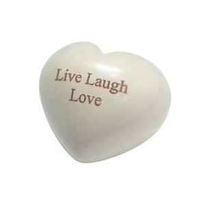 May include: A smooth, off-white heart-shaped stone object featuring the text Live Laugh Love engraved in a brown serif font on its front surface. The object has a polished, rounded appearance with a consistent light cream color throughout.
