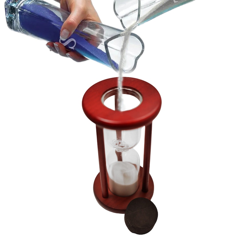 K&ouml;nnte beinhalten: Eine rote Sanduhr aus Holz mit einer Glasbirne, die mit wei&szlig;em Sand gef&uuml;llt ist. Der Sand wird aus einer durchsichtigen Glasflasche mit blauem Sand gegossen.