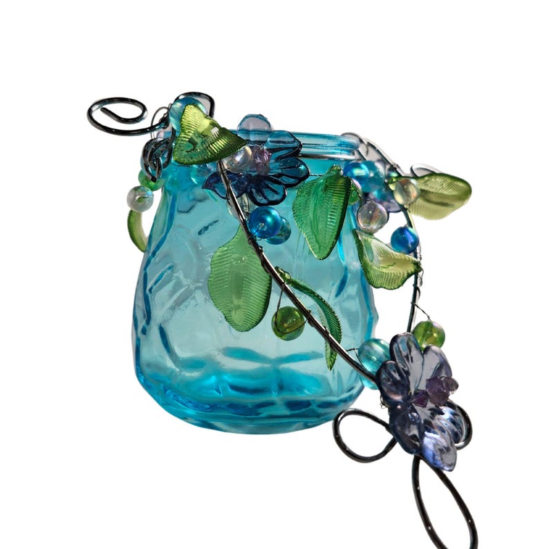 Pu&ograve; includere: Un piccolo vaso di vetro turchese strutturato con un tralcio decorativo in filo di ferro. Il tralcio presenta foglie verdi, perline blu e trasparenti e accenti floreali blu e viola. Il vaso ha un manico decorativo in filo di ferro.