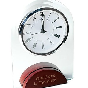May include: A glass clock with a silver face and Roman numeral markings. The clock is on a wooden base with the inscription "Our Love Is Timeless".