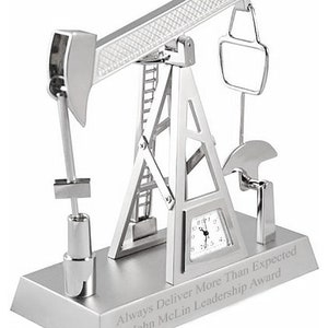 May include: Silver-toned desk clock shaped like an oil derrick with a clock face and a small ladder. The base of the clock is engraved with the text "Always Deliver More Than Expected John McLin Leadership Award".