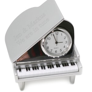 May include: A silver-colored piano-shaped clock with a white face and black hands. The clock is open and the top of the piano has the inscription "Jen & Markus We are in tune".