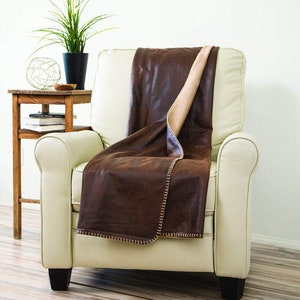 May include: A brown faux leather throw blanket with a tan fleece lining is draped over a white armchair. The blanket has a decorative stitch around the edges.