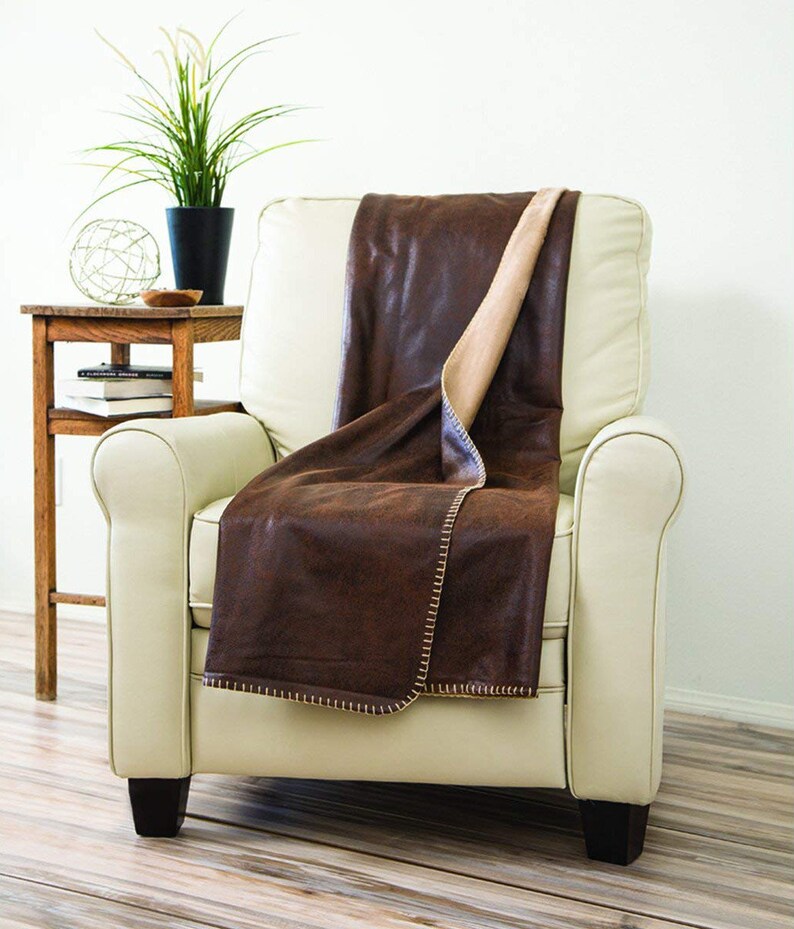 May include: A brown faux leather throw blanket with a tan fleece lining is draped over a white armchair. The blanket has a decorative stitch around the edges.