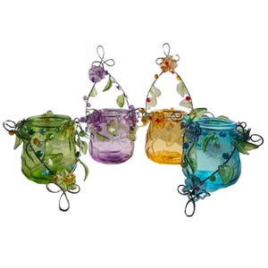 Pu&ograve; includere: Quattro portacandele in vetro colorato con manici in filo metallico e decorazioni floreali. I portacandele sono nelle tonalit&agrave; del verde, viola, giallo e blu. Ogni portacandele ha un design unico con dettagli di perline e foglie decorative.