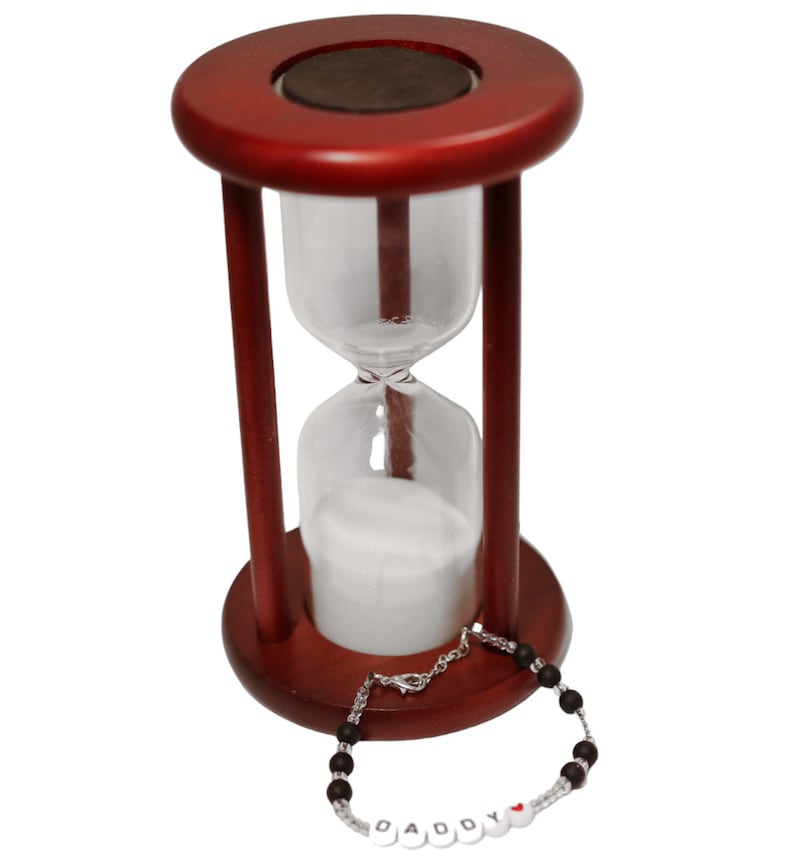 K&ouml;nnte beinhalten: Eine Sanduhr aus Holz mit wei&szlig;em Sand und einem schwarzen Perlenarmband mit dem Wort "DADDY" in wei&szlig;en Perlen mit einer roten Herzperle.