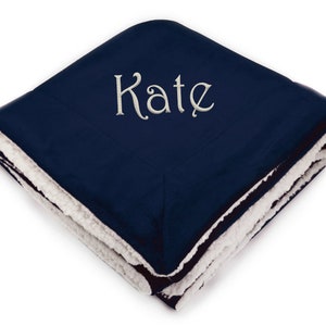 May include: A navy blue fleece blanket with a white sherpa lining. The blanket is folded and the name "Kate" is embroidered on the front in a white cursive font.