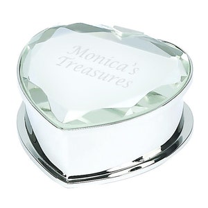 May include: A heart-shaped trinket box with a clear, faceted lid and a shiny silver base. The lid is engraved with the words "Monica's Treasures" in a delicate script. The box is designed to hold small jewelry items or keepsakes.