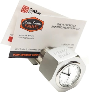 May include: A silver metal desk clock shaped like a large nut with a clock face and the words "Johnny Brush" engraved on the side. The clock is sitting on top of a business card that reads "Dunn-Edwards Paints" and "The #1 Choice of Painting Professionals!"