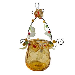 Pu&ograve; includere: Un portacandele decorativo in vetro ambrato con superficie strutturata. Il supporto &egrave; adornato con una struttura metallica, fiori, perline e dettagli a forma di foglie. Ha un manico in filo metallico per appenderlo.