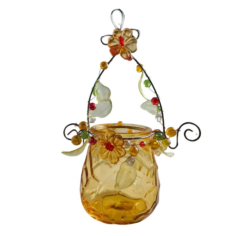 Pu&ograve; includere: Un portacandele decorativo in vetro ambrato con superficie strutturata. Il supporto &egrave; adornato con una struttura metallica, fiori, perline e dettagli a forma di foglie. Ha un manico in filo metallico per appenderlo.