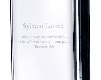 Può includere: Un blocco di vetro rettangolare trasparente con il nome "Sylvain Lavoie" e un versetto biblico inciso. Il blocco ha un lato scuro e riflettente, probabilmente un oggetto decorativo o un premio.