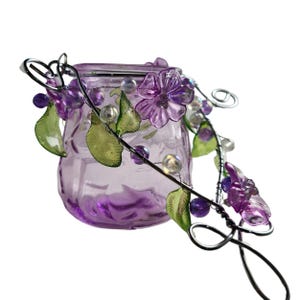 Pu&ograve; includere: Un piccolo vaso di vetro viola con un tralcio decorativo in filo metallico con fiori viola, foglie verdi e perline trasparenti. Il vaso ha una superficie strutturata ed &egrave; destinato a scopi decorativi.