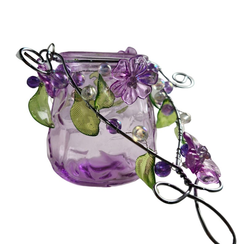 Pu&ograve; includere: Un piccolo vaso di vetro viola con un tralcio decorativo in filo metallico con fiori viola, foglie verdi e perline trasparenti. Il vaso ha una superficie strutturata ed &egrave; destinato a scopi decorativi.