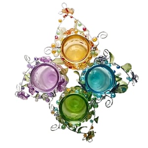 Pu&ograve; includere: Un portacandele decorativo con quattro bicchieri di vetro colorati in giallo, viola, turchese e verde. I bicchieri sono circondati da una cornice in filo d'argento con accenti floreali e perlati. Il portacandele &egrave; a forma di stella.