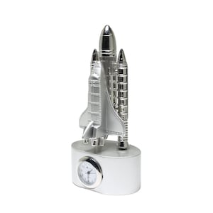 May include: Silver-colored space shuttle figurine with a clock. The clock has a white face with black numbers and hands. The shuttle is detailed with wings, boosters, and a nose cone. The base is silver.