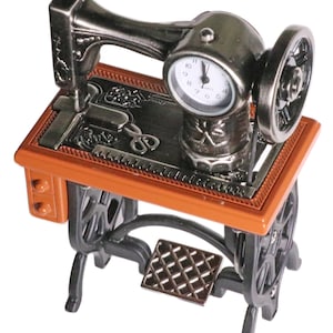 Peut inclure: Une horloge miniature en forme de machine &agrave; coudre de couleur argent&eacute;e avec une base brune et des pieds noirs. Le cadran de l'horloge a des aiguilles noires et des chiffres noirs.