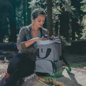 May include: A gray backpack cooler with black straps and a black zipper. The cooler is filled with bottles and is sitting on a tree stump in a forest setting.