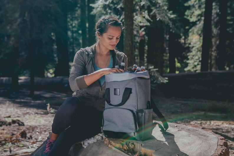 May include: A gray backpack cooler with black straps and a black zipper. The cooler is filled with bottles and is sitting on a tree stump in a forest setting.