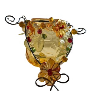 Pu&ograve; includere: Un portacandele decorativo in vetro di colore giallo-arancio. Il portacandele &egrave; adornato con decorazioni floreali e a forma di foglie, oltre a perline colorate. La struttura metallica presenta volute decorative e sostiene il contenitore di vetro.