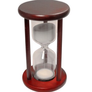 K&ouml;nnte beinhalten: Eine Sanduhr aus Holz mit einer durchsichtigen Glasbirne, die mit wei&szlig;em Sand gef&uuml;llt ist. Die Sanduhr steht aufrecht auf einem wei&szlig;en Hintergrund.