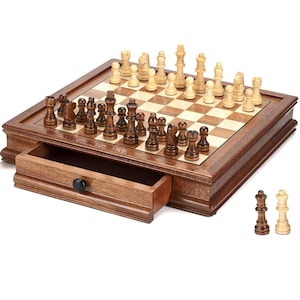 May include: A wooden chess set with a square board featuring alternating light and dark wood squares. The board includes a pull-out storage drawer with the engraved text Our Greatest Game Is Life Together. Dark and light wooden chess pieces are arranged.