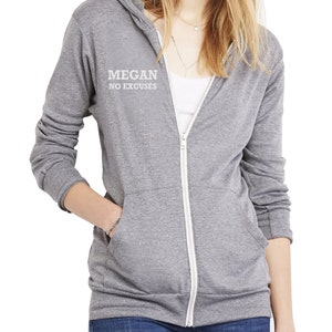 May include: A heather gray zip-up hoodie with a white zipper and pockets. The hoodie features the text "MEGAN NO EXCUSES" in white on the upper left chest. The model is wearing blue jeans and a white tank top underneath.